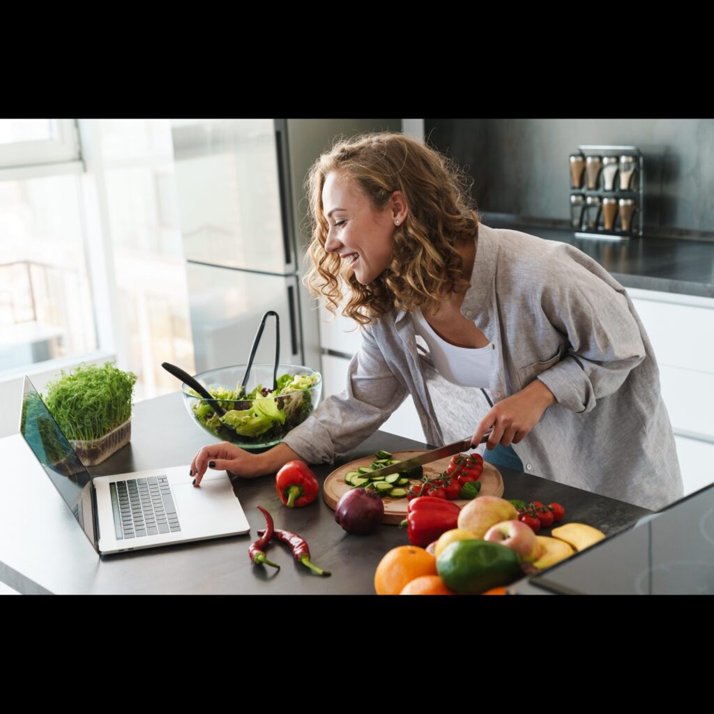 Happy young woman making a salad at the kitchen, chopping vegetables, looking at laptop computer